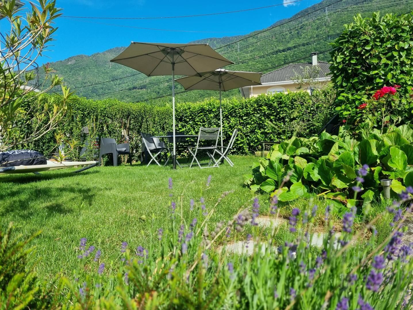 Un jardin avec une pelouse, une table entourée de chaises, un grand parasol et un espace planté de différentes espèces végétales.