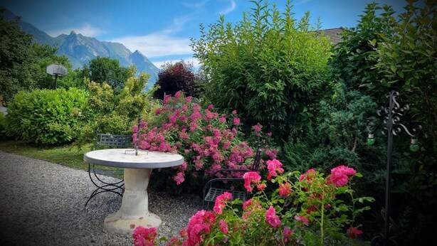 En trädgård med ett runt bord och stolar, omgiven av frodig grönska och rosa blommor.