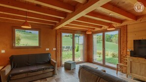 A cozy living room with a sofa, a TV, and large windows offering a view of the outdoors. - LE Chalet DE LA Source (MONTAGNOLE)