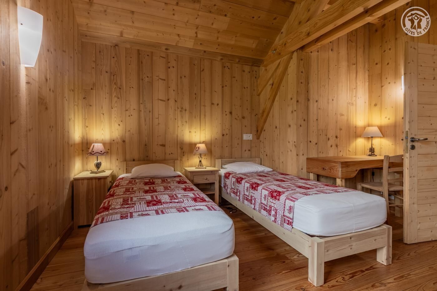 Una habitación de cabaña de madera con dos camas, cada una con colcha de rayas rojas y blancas, mesitas de noche de madera con lámparas y un escritorio de madera con una silla.