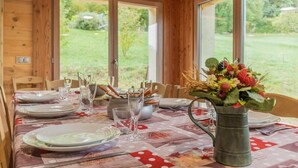 A dining table set for four with a floral tablecloth, a vase of flowers, and a view of the outdoors through large windows. - LE Chalet DE LA Source (MONTAGNOLE)