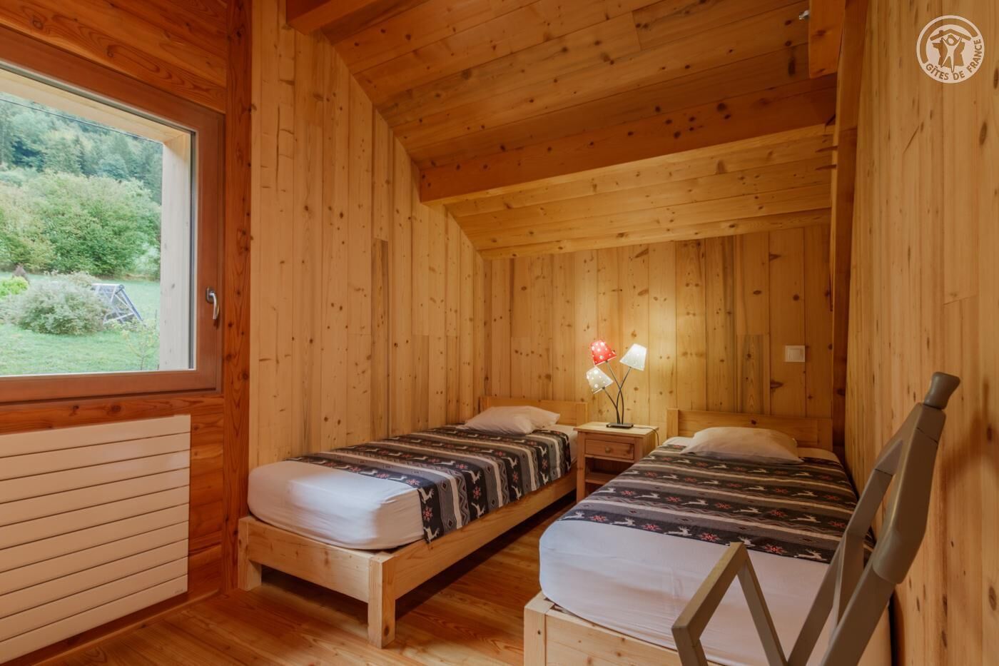 Habitación de cabaña de madera con dos camas, una mesita de noche, una lámpara y ventana con vistas a la vegetación.