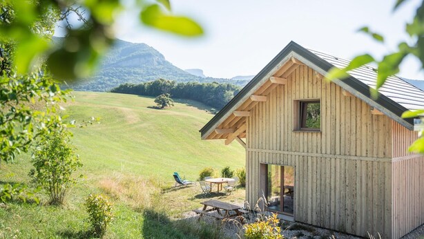 A wooden cabin with a sloped roof, a small window, and a door. It is situated in a grassy field with a mountain in the background. - LE Chalet DE LA Source (MONTAGNOLE)