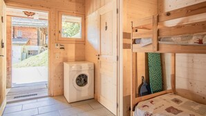 Een houten hut met een wasmachine, een stapelbed en een raam met uitzicht op een huis en groen.