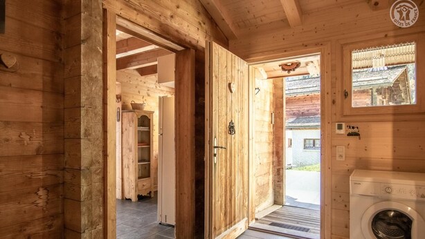Interieur van een houten hutje met een wasmachine, een klein krukje en een deur naar buiten.