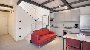 A modern kitchen with white cabinets, a stainless steel refrigerator, and a microwave. There is a red sofa and a dining table with chairs. A staircase with a metal railing leads to another room. - Frantoio a Monte-Apartment near the lake (Malcesine)