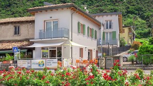 A two-story building with a balcony, a signboard advertising beach-related services, and a lush green hillside in the background. - Frantoio a Monte-Apartment near the lake (Malcesine)