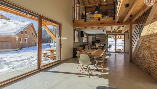 A spacious dining area with a large table and chairs, a kitchen in the background, and a view of a snowy landscape through large windows. - LE Davo - Hauteluce - Gîte (HAUTELUCE)