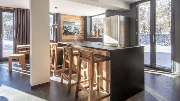 A modern kitchen with a central island, wooden bar stools, and large windows offering a view of the outdoors.
