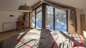 A bedroom with a large window, a wooden chair, a table, and a hanging light fixture.