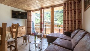 A cozy living room with a wooden ceiling, a sofa, a TV, and a view of a garden through large windows. - LES Lauzes 1 - Bonneval SUR ARC (BONNEVAL SUR ARC)