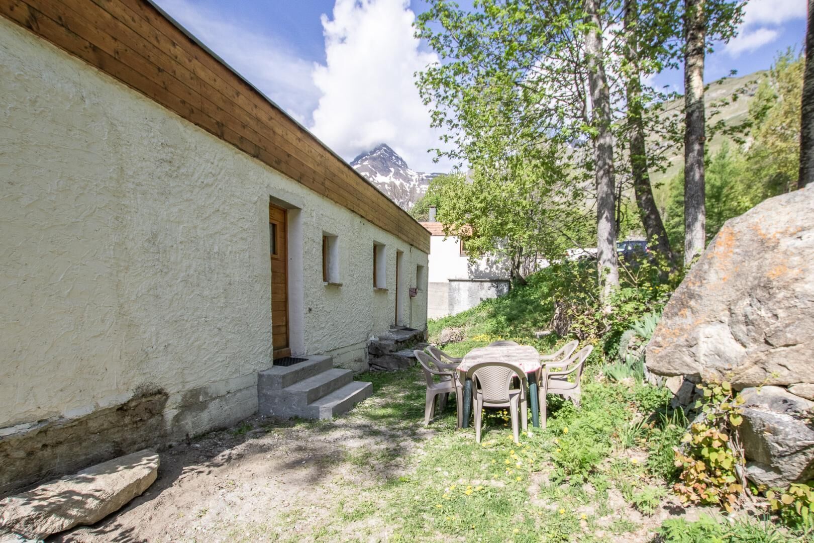 A rustic building with a wooden upper floor and a white lower section, a small outdoor seating area with plastic chairs, and a mountainous backdrop.