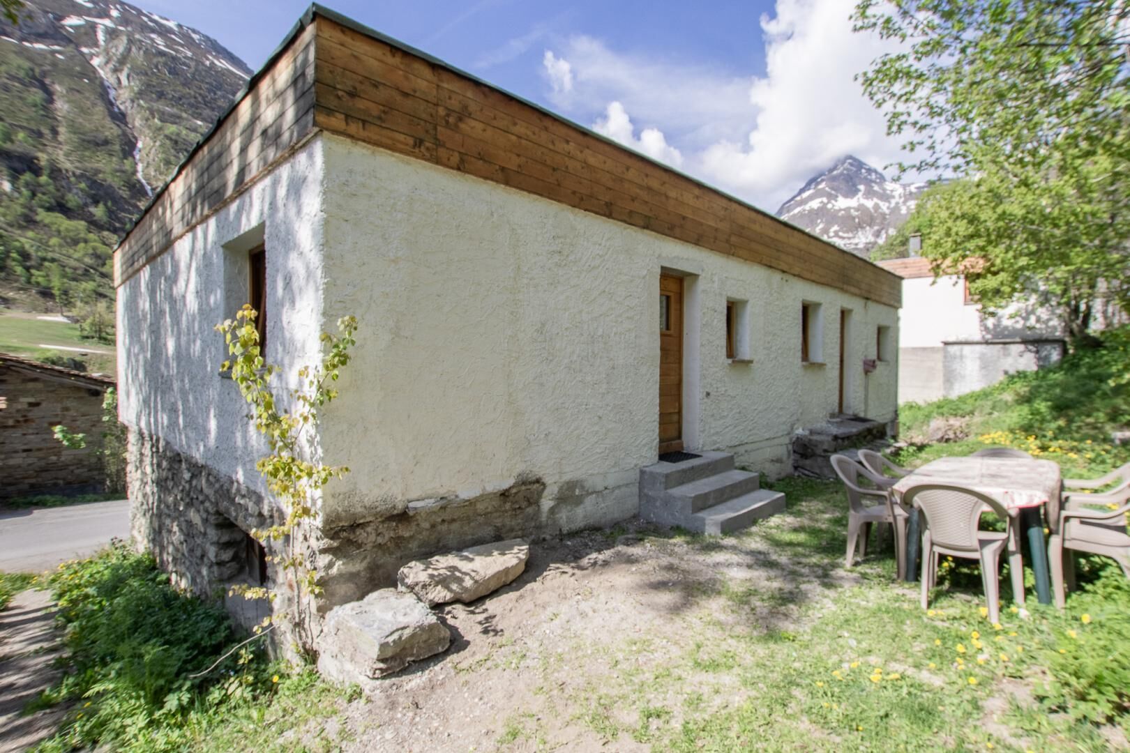 A small, rustic building with a wooden upper floor and a white lower wall, a set of outdoor chairs, and a stone wall nearby.