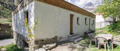 A small, rustic building with a wooden upper floor and a white lower wall, a set of outdoor chairs, and a stone wall nearby.