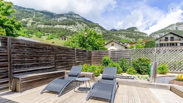 A wooden deck with two lounge chairs and a small table, surrounded by a wooden fence, overlooking a mountainous landscape with houses in the background. - LE Barillon - Avrieux (AVRIEUX)