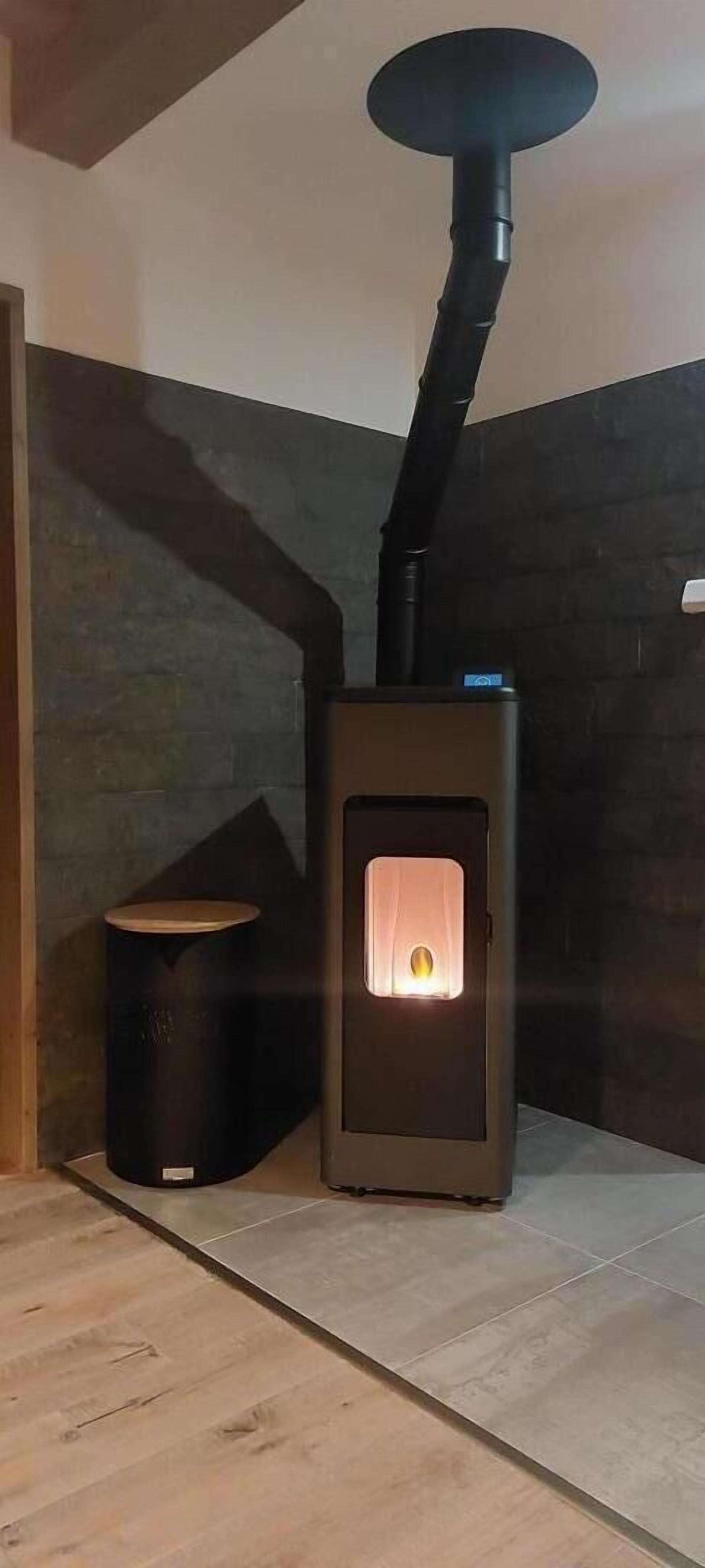 Un poêle à bois avec une porte en verre, un petit tabouret et un mur sombre.