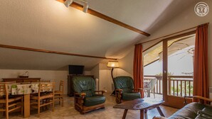 Een ruime woonkamer met een eettafel, twee groene leren fauteuils, een houten salontafel en een bank. Een televisie is aan de muur bevestigd, en er is een balkon met uitzicht.