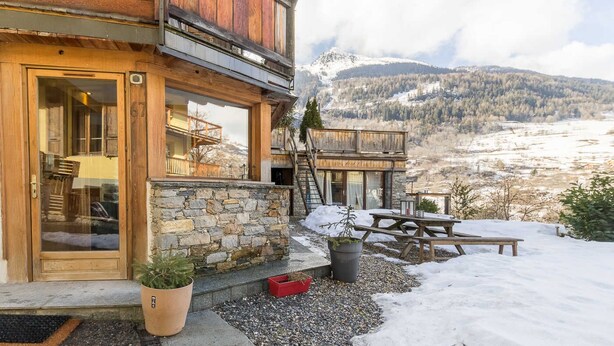 A wooden chalet with a stone foundation, large windows, and a view of a snowy mountain landscape. - Maison Adele - Ste-foy-tarentaise (SAINTE FOY TARENTAISE)