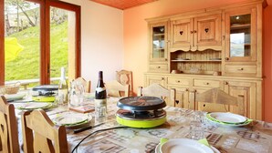 A dining area with a table set for four, wooden chairs, a buffet cabinet, and a view of a green landscape through large windows. - LE Petit Bonheur - St-jean-d'arves (SAINT JEAN D'ARVES)