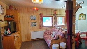 A cozy room with a wooden floor, a sofa with a patterned blanket, a dining table with chairs, a window with curtains, and a wall-mounted deer head decoration.