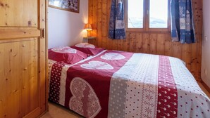 A cozy room with a wooden paneled wall, a bed with a red and white quilt, a nightstand with a lamp, and a window with curtains.