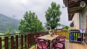 Une terrasse en bois avec une table et des chaises, un barbecue et une vue sur une vallée verdoyante et des montagnes.