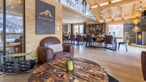 Un intérieur chaleureux avec un plafond en bois, un coin repas, une cheminée et une grande fenêtre donnant sur un paysage enneigé.
