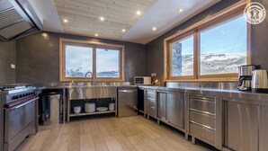 Une cuisine moderne dotée d’appareils électroménagers en acier inoxydable, d’un plafond en bois et de grandes fenêtres offrant une vue sur un paysage enneigé.