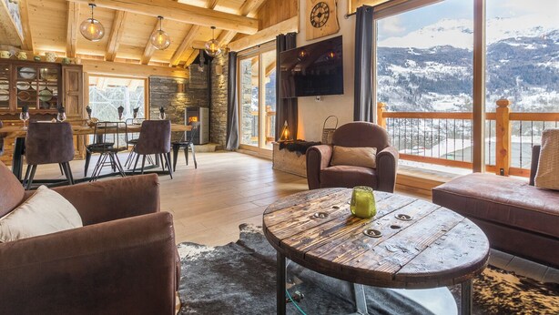 Un vaste espace de vie agrémenté d’une cheminée en pierre, de poutres apparentes en bois et d’une grande fenêtre offrant une vue sur les montagnes.