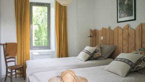 A bedroom with two beds, a wooden headboard, a window with curtains, a chair, and a hat on the bed. - Gite DU Miron - St-paul-sur-yenne (ST-PAUL-SUR-YENNE)