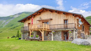 Un chalet en bois doté d’un balcon, avec des fondations en pierre et un paysage montagneux en arrière-plan.