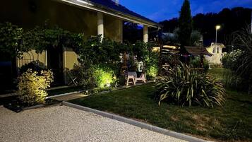 Un bâtiment doté d’un espace extérieur aménagé avec une zone de repos couverte, d’une allée en gravier et de diverses plantes.