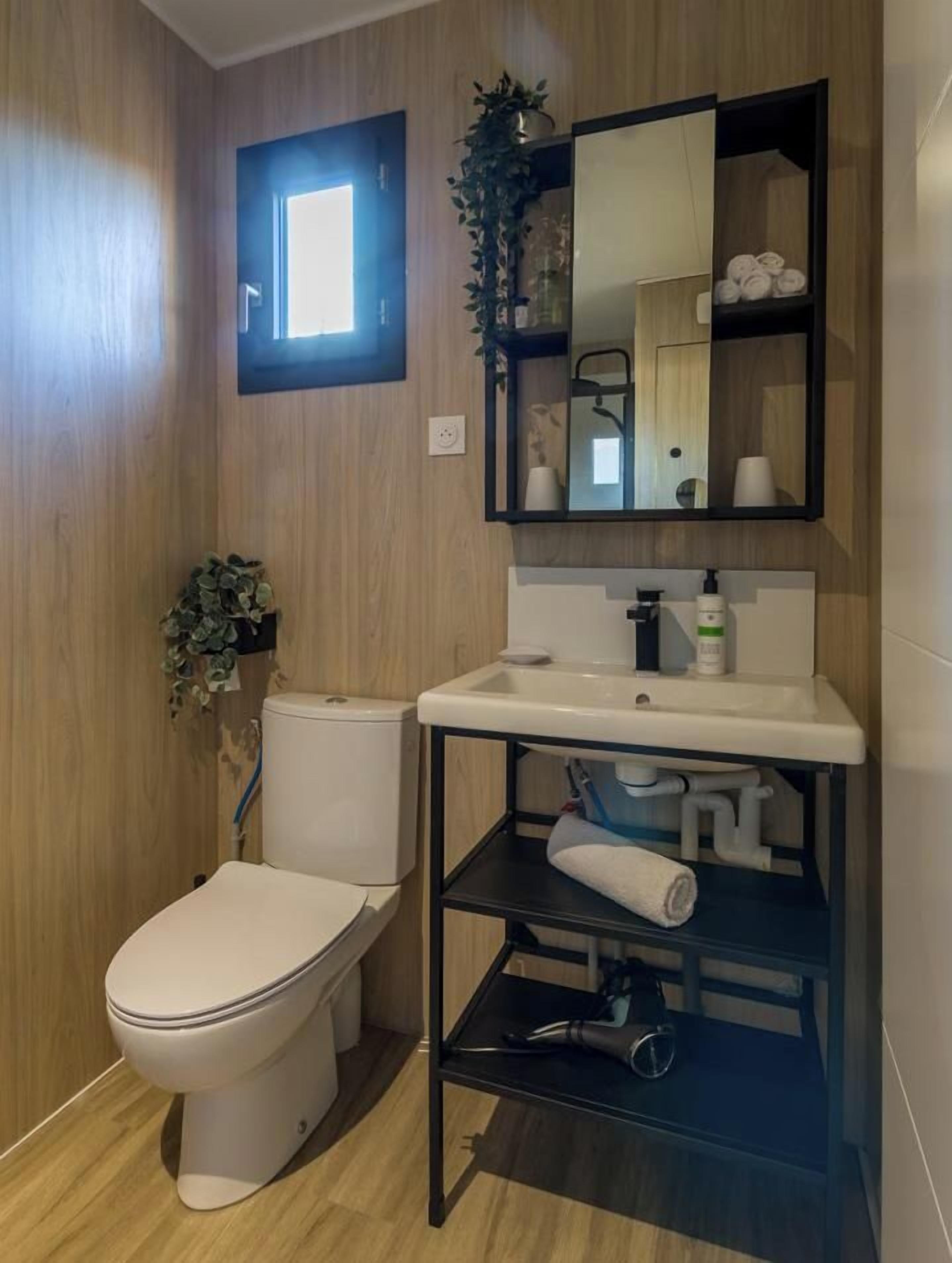 A modern bathroom with a wooden wall, a white toilet, a sink with a mirror above it, and a shelf with toiletries.