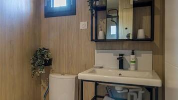 A modern bathroom with a wooden wall, a white toilet, a sink with a mirror above it, and a shelf with toiletries.