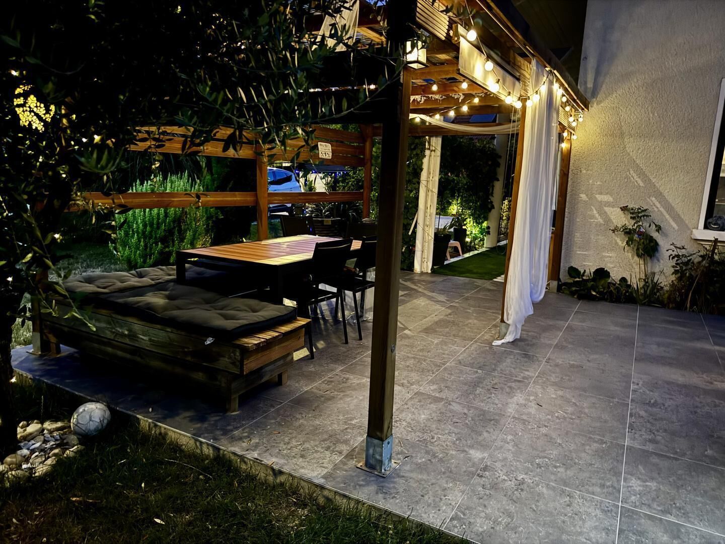 An outdoor patio area with a wooden table and chairs, a cushioned bench, and string lights.