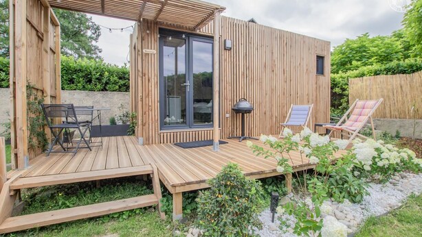 Une terrasse en bois avec des meubles d’extérieur, une pergola et un petit jardin agrémenté de fleurs blanches et de rochers.