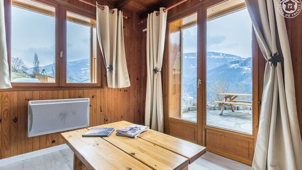 A wooden room with a table, two windows with curtains, a radiator, and a view of mountains.