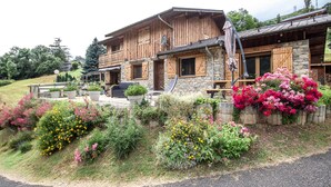 A rustic wooden house with stone walls, a balcony, and a garden with various flowers.