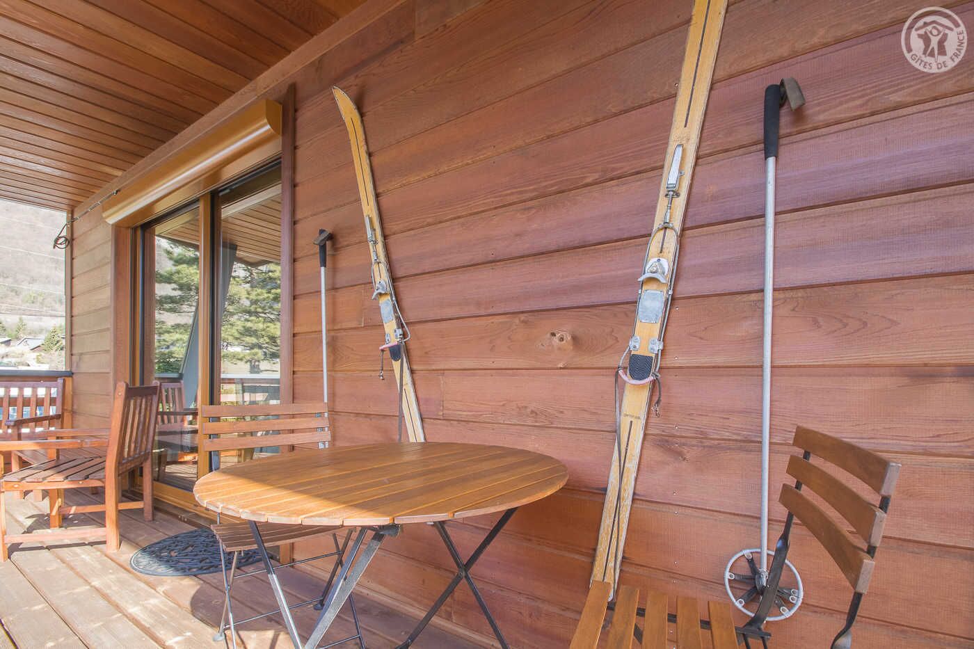 Een houten chalet met een tafel en stoelen op het terras, ski-uitrusting aan de wand en een deur die naar buiten leidt.