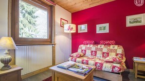Una habitación acogedora con una pared roja, techo de madera, ventana con vista a árboles, sofá acolchado, una lámpara y una mesa con libros.