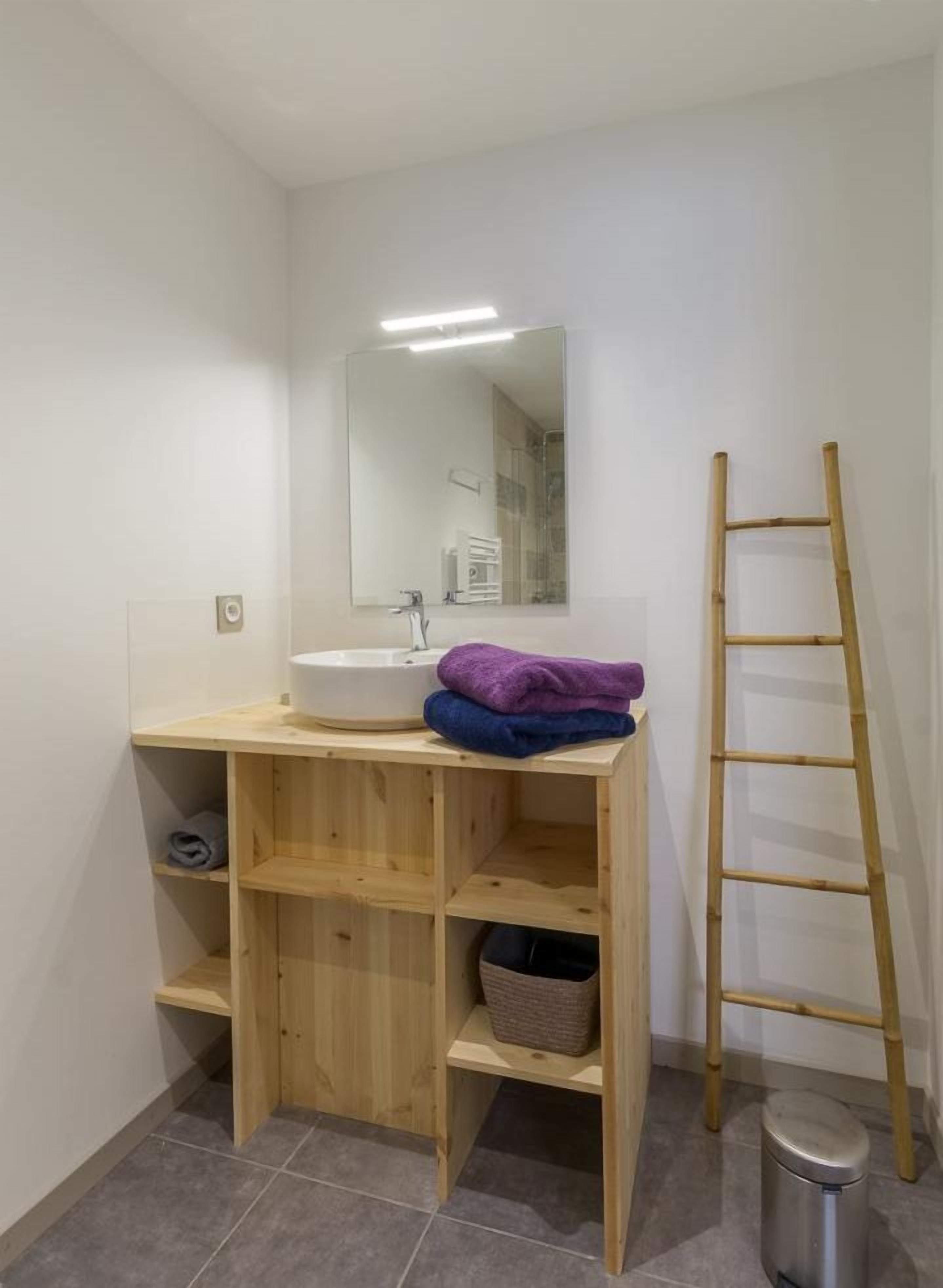Une salle de bain avec un meuble-lavabo en bois, un lavabo blanc, un miroir et un escabeau en bambou.