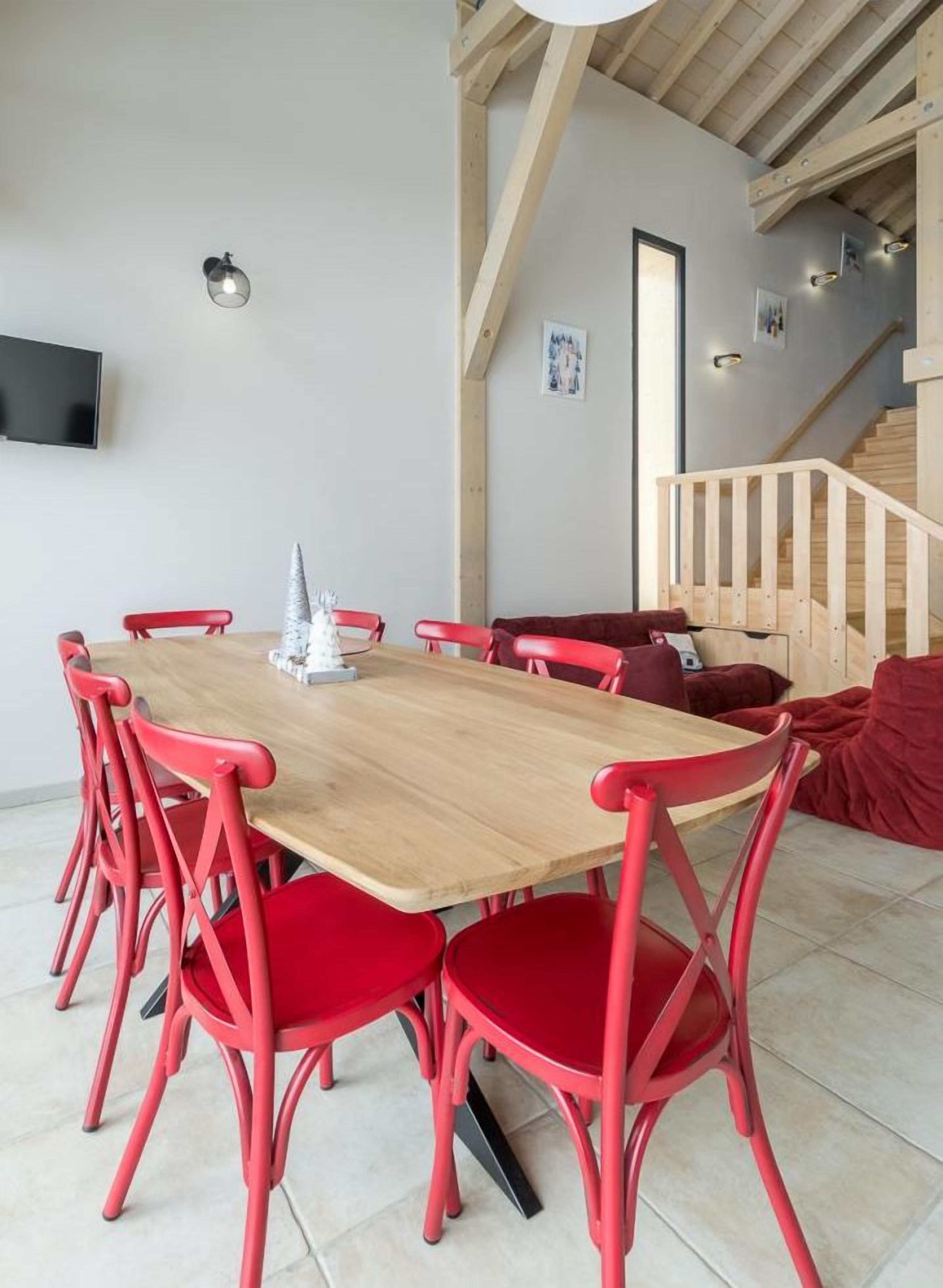 Un área de comedor con una mesa de madera y sillas rojas, un televisor montado en la pared y una escalera al fondo.