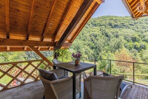 Eine Holzterrasse mit Korbmöbeln und einem Tisch, mit Blick auf einen bewaldeten Hang.