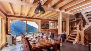 Un comedor de madera con una mesa grande y sillas, una escalera y vistas a montañas.