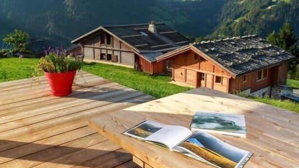 Una terraza de madera con un banco y una mesa, una planta en maceta roja, y una cabaña de madera con un paisaje montañoso al fondo.