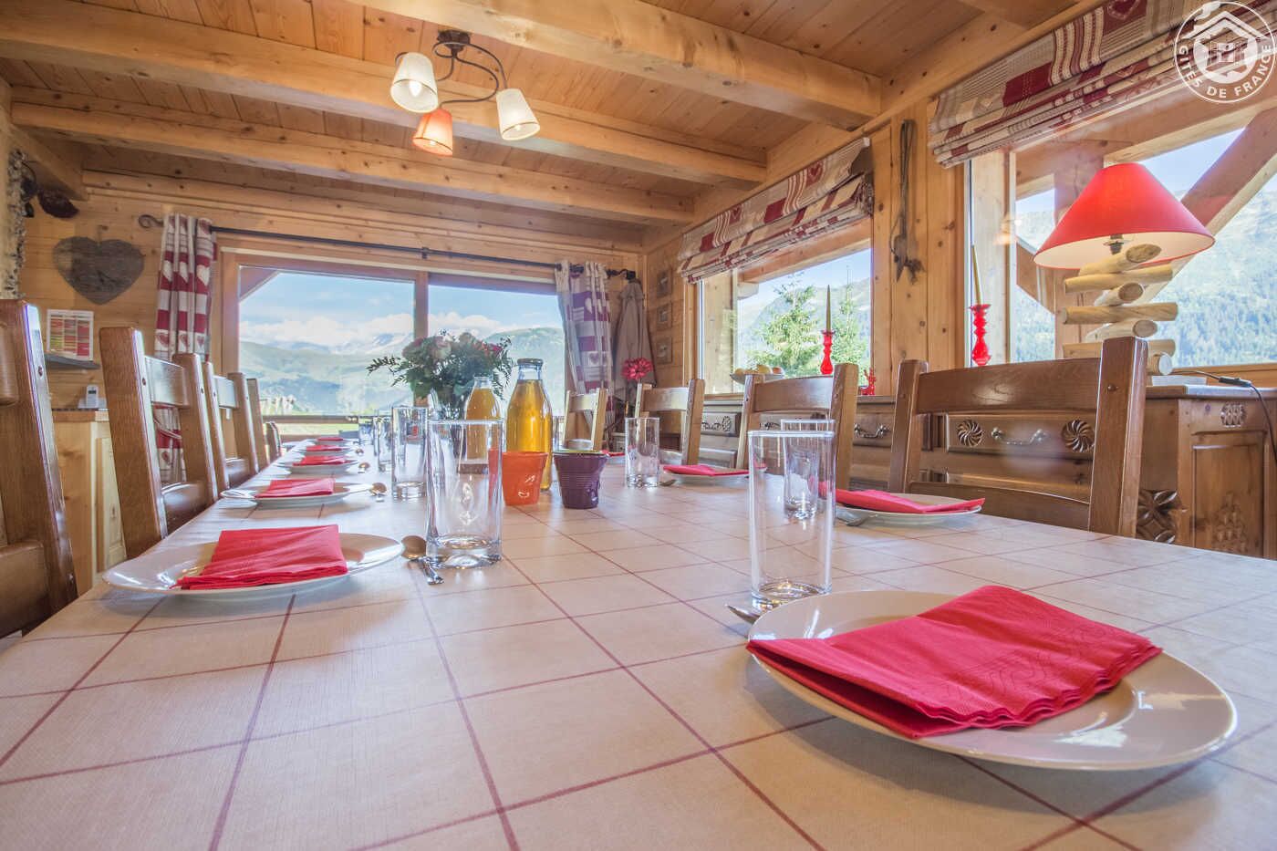 Uma sala de jantar com uma mesa comprida posta para refeição, paredes de madeira e janelas amplas com vista para o exterior.