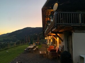 A wooden building with a balcony, outdoor seating, and a view of the hills.