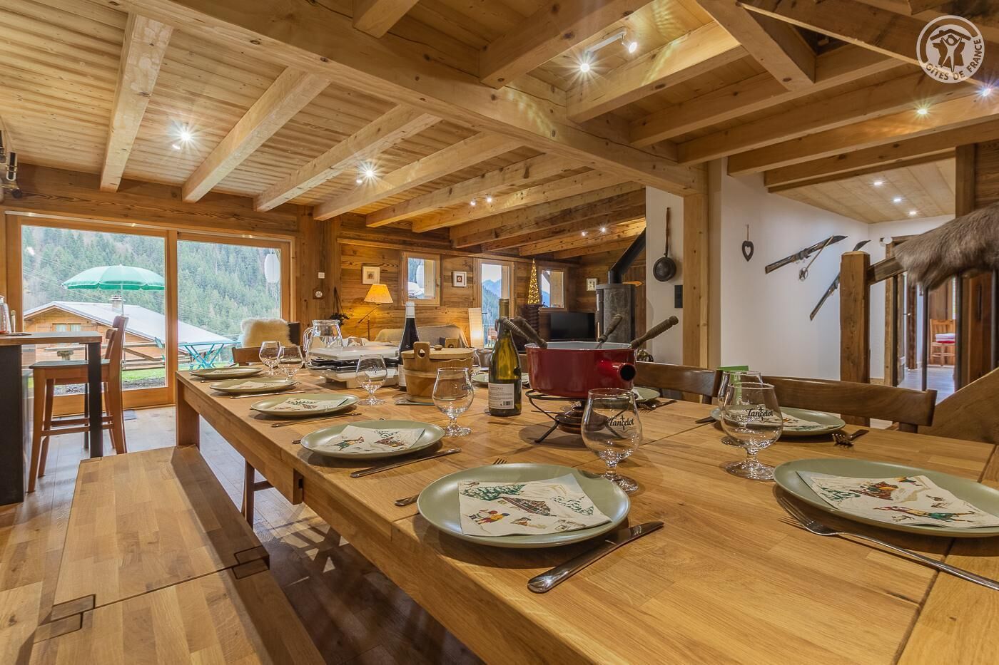 Un coin repas en bois, avec une longue table dressée pour le repas, une cheminée et une vue sur l’extérieur.