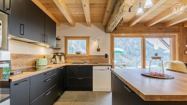 Una cocina moderna con armarios oscuros, encimera de madera, lavavajillas y vistas a montañas nevadas a través de amplios ventanales.