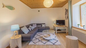 Un salon chaleureux avec un plafond en bois, un canapé gris, un téléviseur et une fenêtre ornée de plantes.